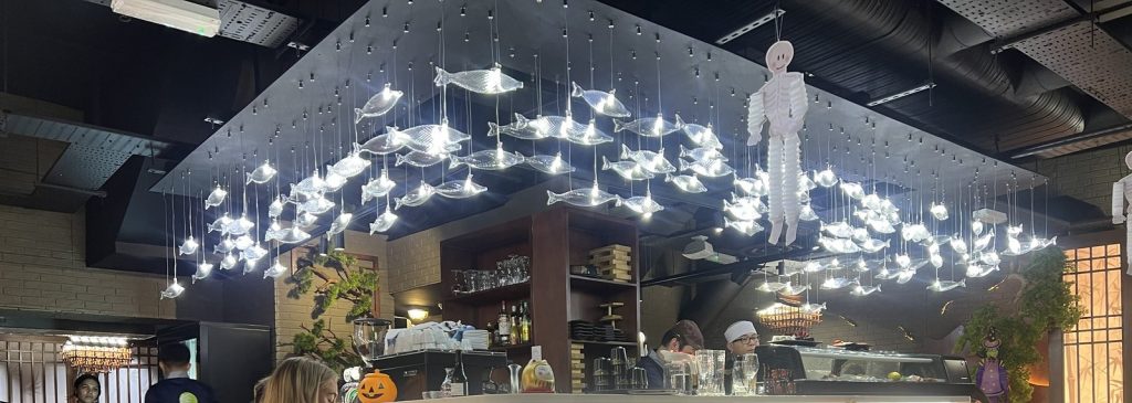 A ceiling of hanging glass fish lights above a sushi counter, with a chef at work behind it