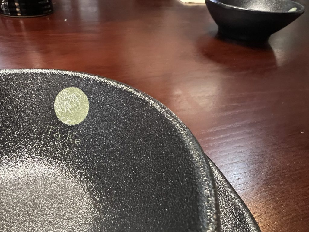 Close-up of a dark textured dish with a small green Ta Ke logo, on a wooden table