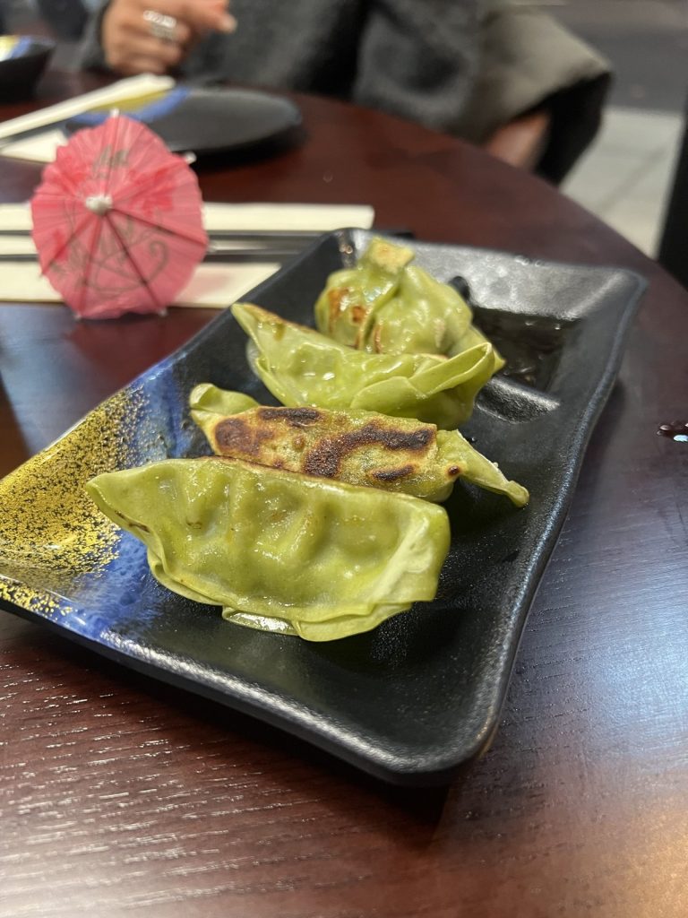 Four green vegetable gyoza on a black and blue plate, with a paper parasol on the table behind