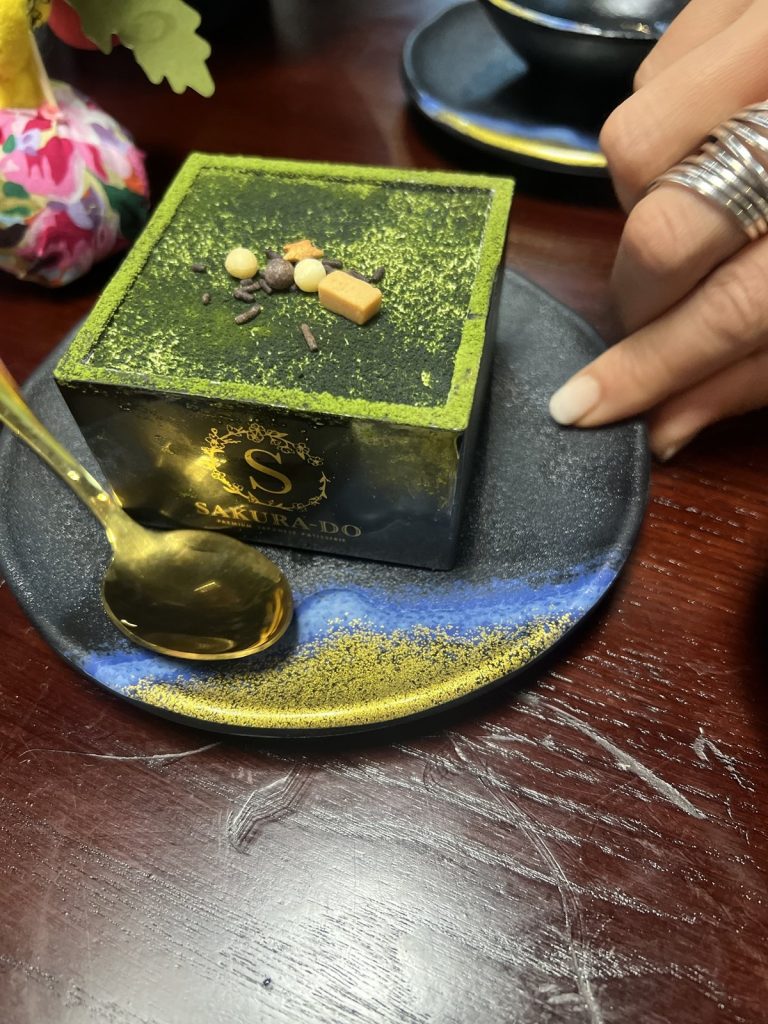 A square matcha tiramisu dusted with green powder, in a black box branded Sakura-Do, with a gold spoon