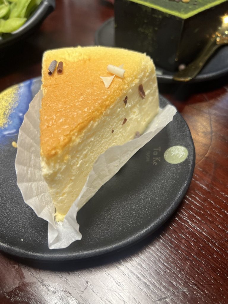 A wedge of pale, fluffy Japanese cheesecake in a paper case on a Ta Ke plate