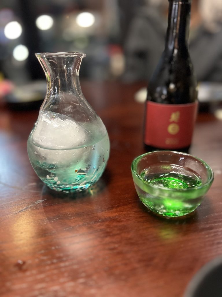 A glass sake carafe with ice, a small green glass cup and a bottle with a red label