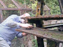A man in a striped shirt bends over the Merton Abbey Mills waterwheel, working on one of its wooden paddles with a hand tool