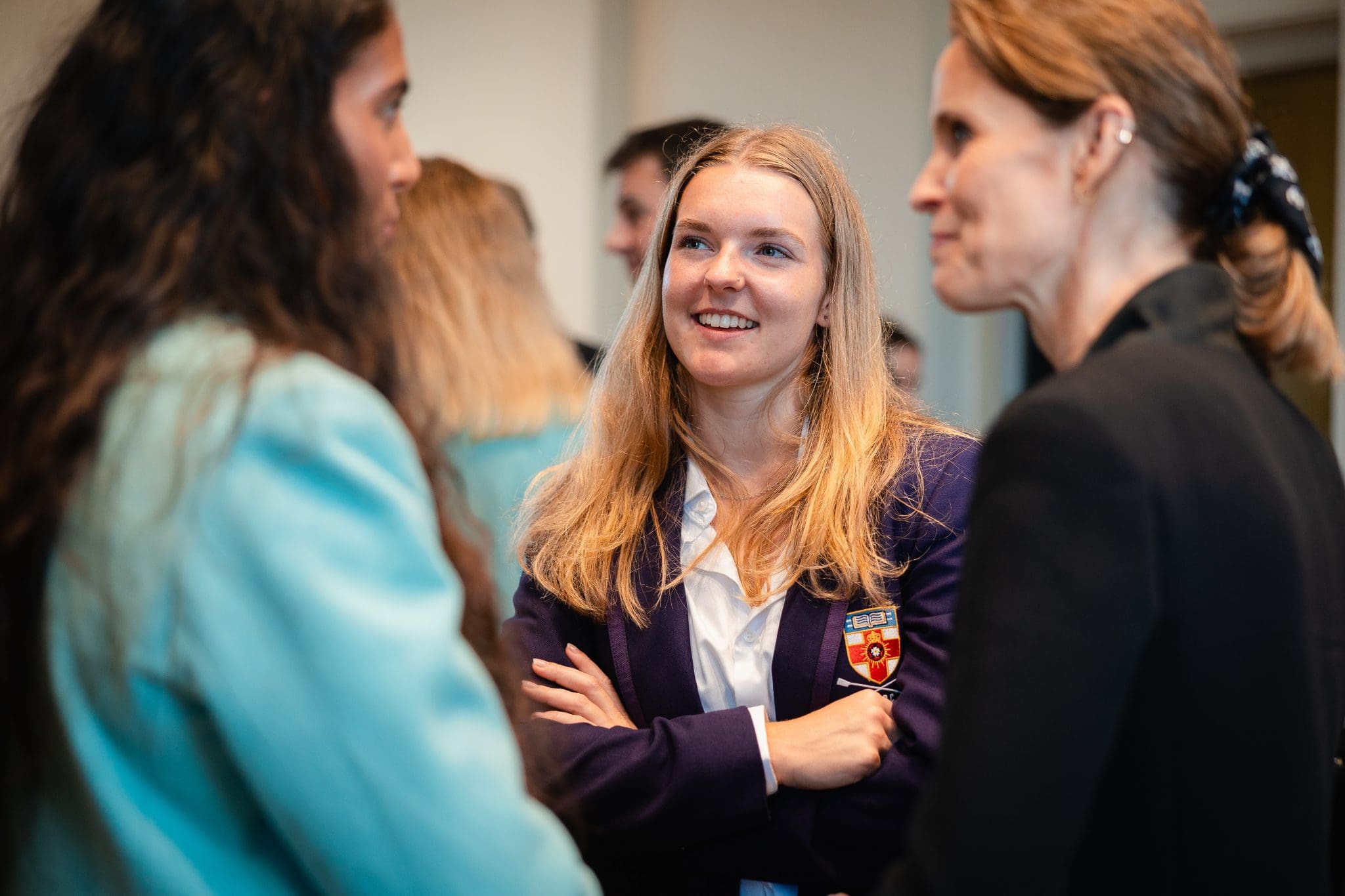 A smiling rower in a dark blue blazer with a crest on the pocket, arms folded, talks with two women at the Presidents' Challenge