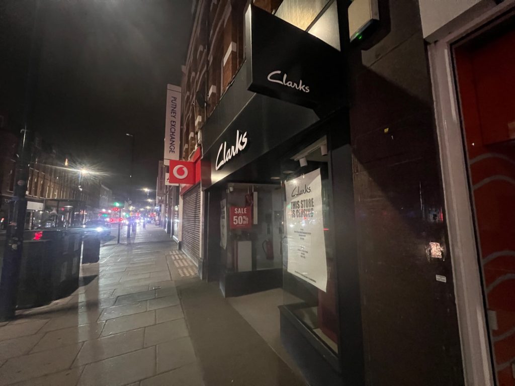 Night view along Putney High Street past Clarks, with its closing poster and sale sign, towards the Putney Exchange sign