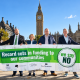 Four people hold a green banner reading "Record cuts in funding to our communities, we say NO" in Parliament Square, with Big Ben behind