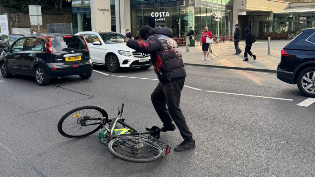 A man in a dark padded jacket stands over a bicycle lying in the road on Upper Richmond Road, with stopped cars beside him and Costa Coffee at Stamford Square behind