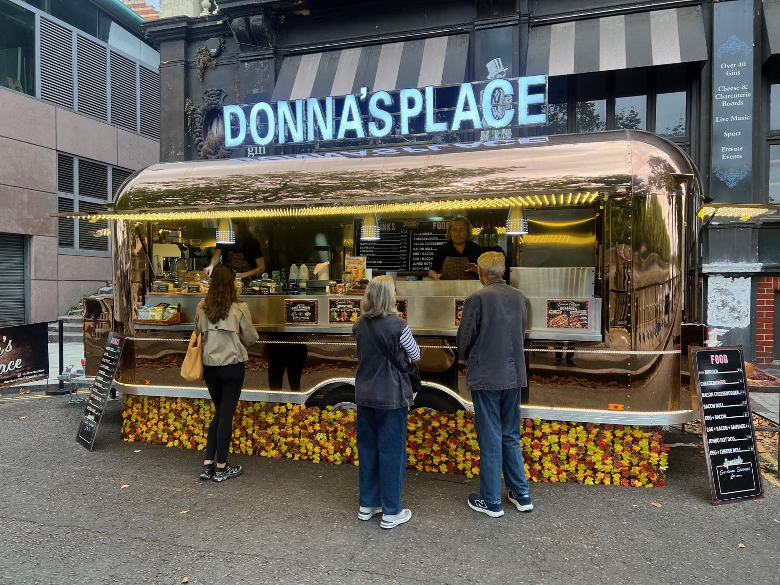 Three customers, seen from behind, at the counter of a gold-toned mirror-finish trailer with lit Donna's Place lettering, while staff serve inside