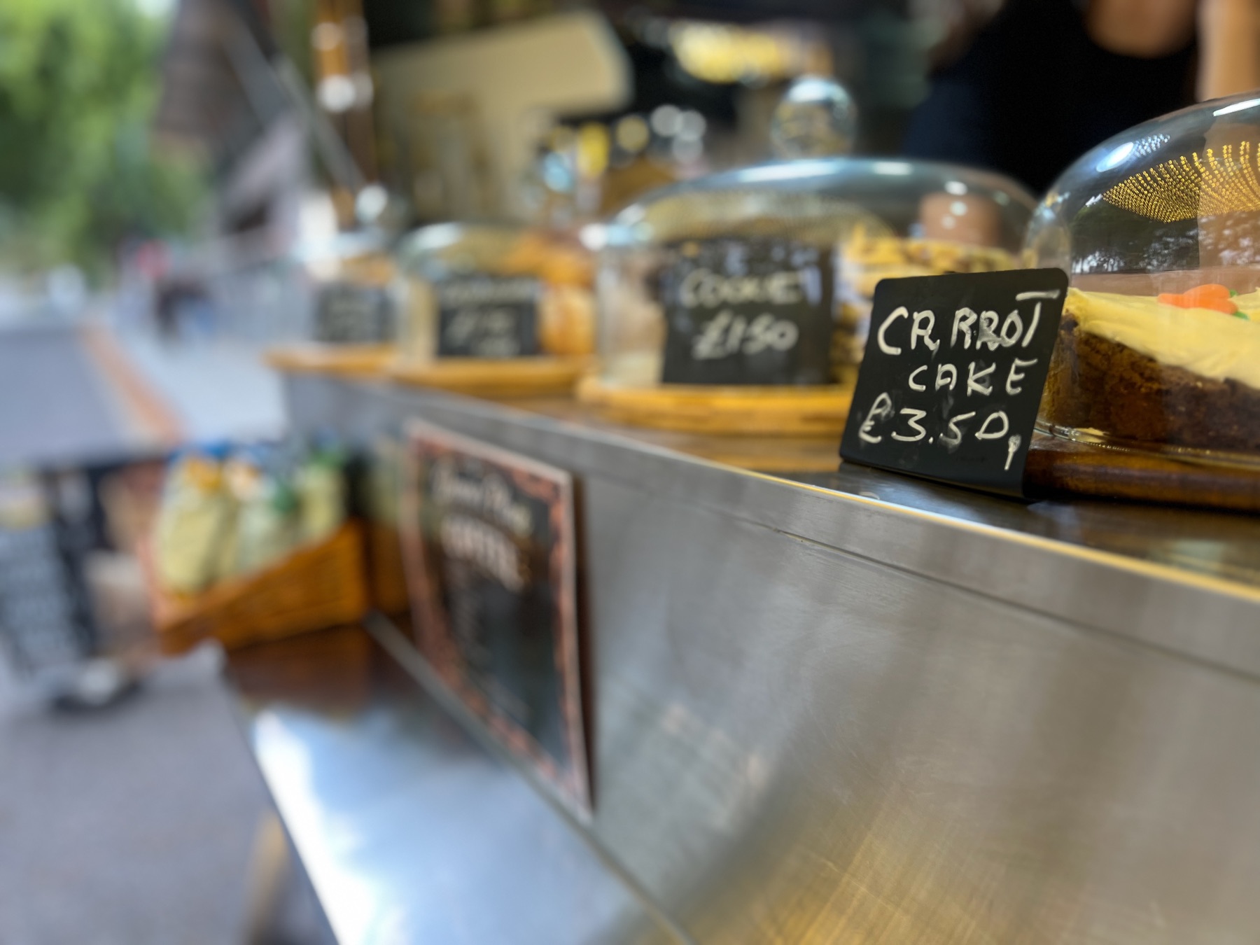 Cakes under glass domes on a steel counter, with handwritten chalk tags