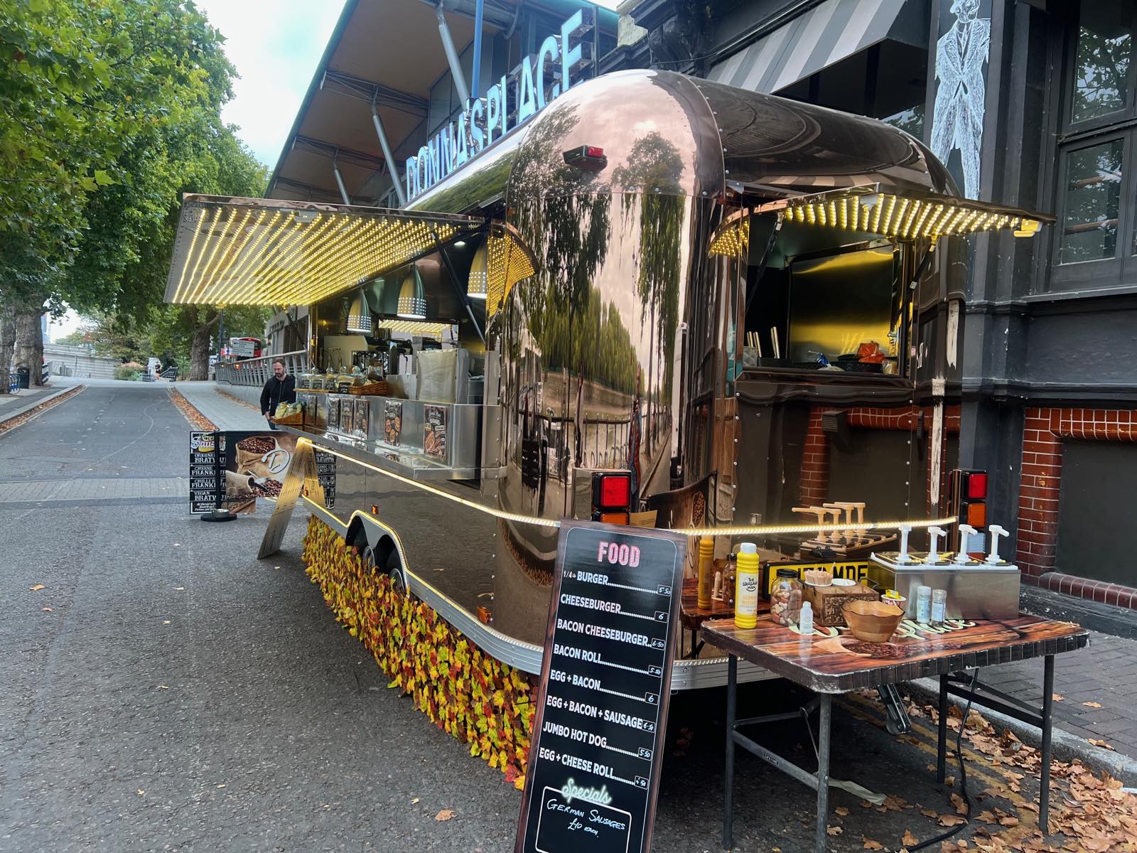 Side view of a curved mirror-finish trailer with two lit awnings open, parked beside the tree-lined embankment