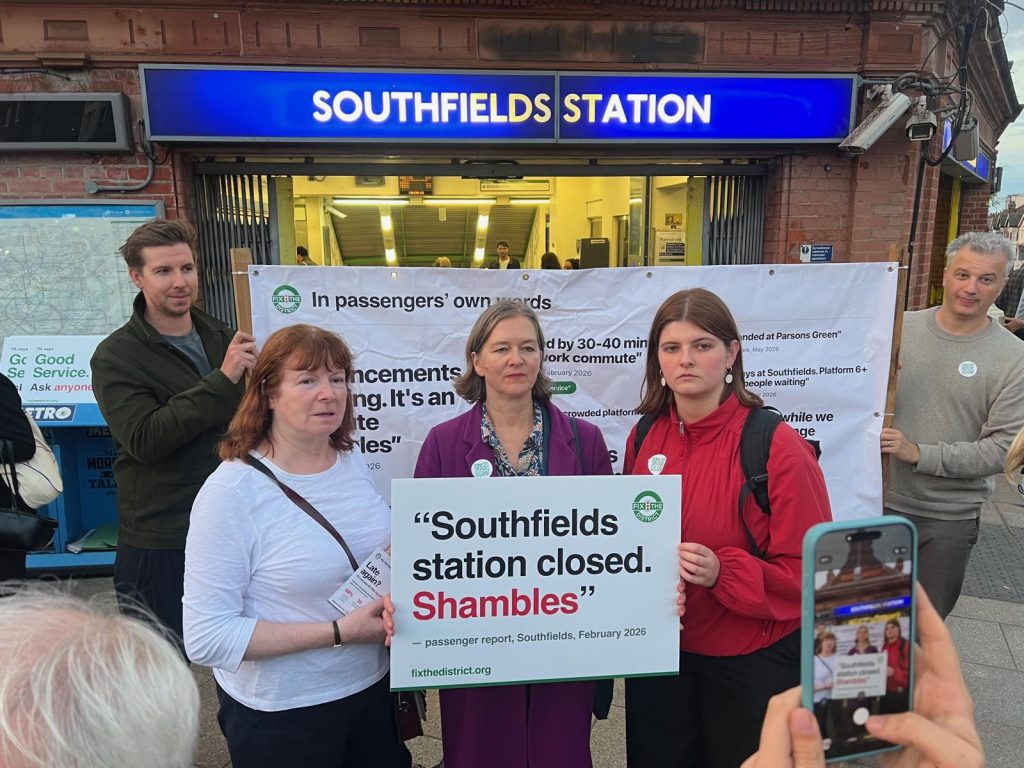 Councillor Angela Ireland, Fleur Anderson MP and councillor Jasmin Glynne with a Fix the District placard reading Southfields station closed. Shambles