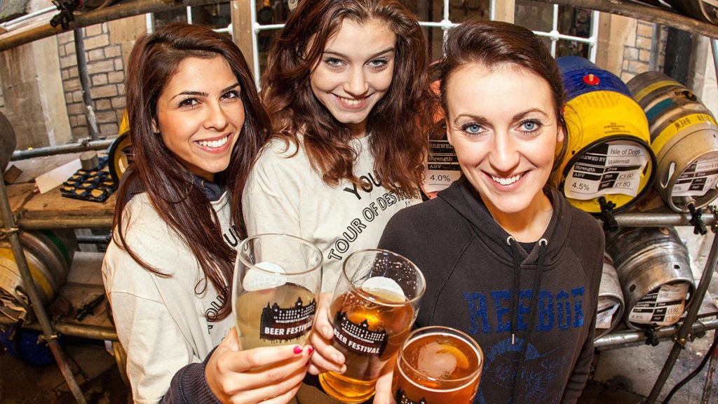 Three women raise festival glasses of beer in front of racked casks at Le Gothique