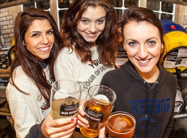 Three women raise festival glasses of beer in front of racked casks at Le Gothique