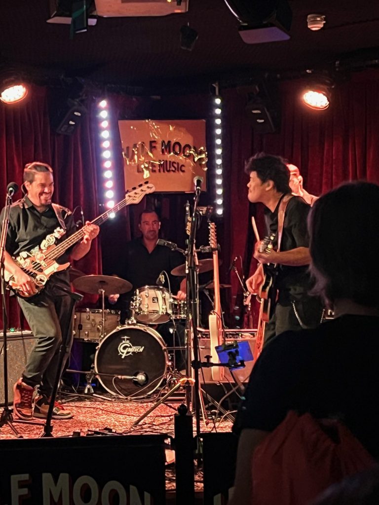 Paranohead’s bassist laughing on stage as a guitarist turns towards him, with the drummer behind.