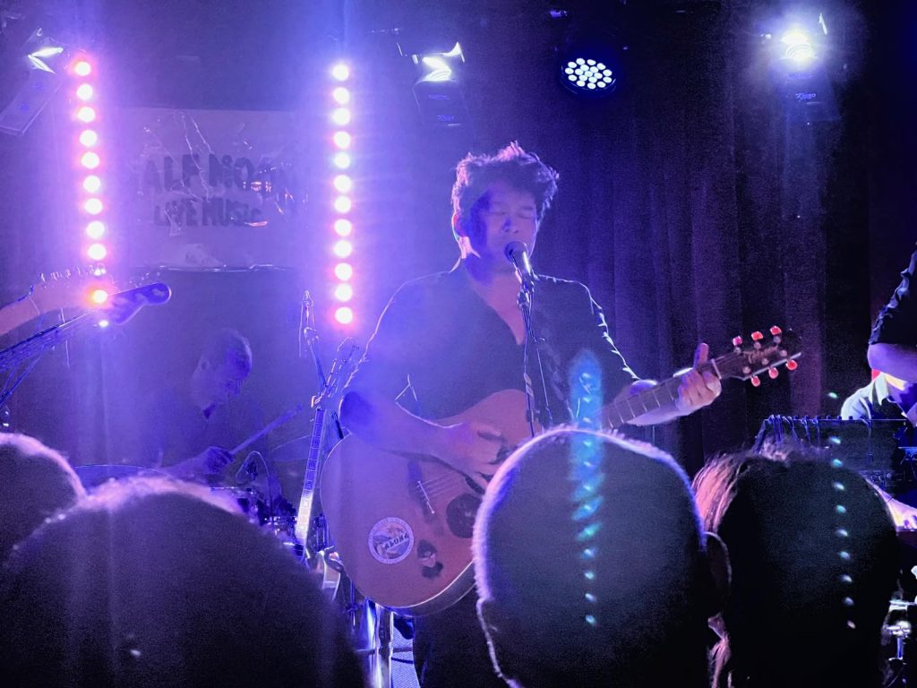 Robb Tito singing with his eyes closed and playing acoustic guitar in blue and white stage light.