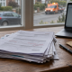 Stack of printed forms with red pen alterations on a desk beside a laptop, with a parked car and recovery van visible through the window.