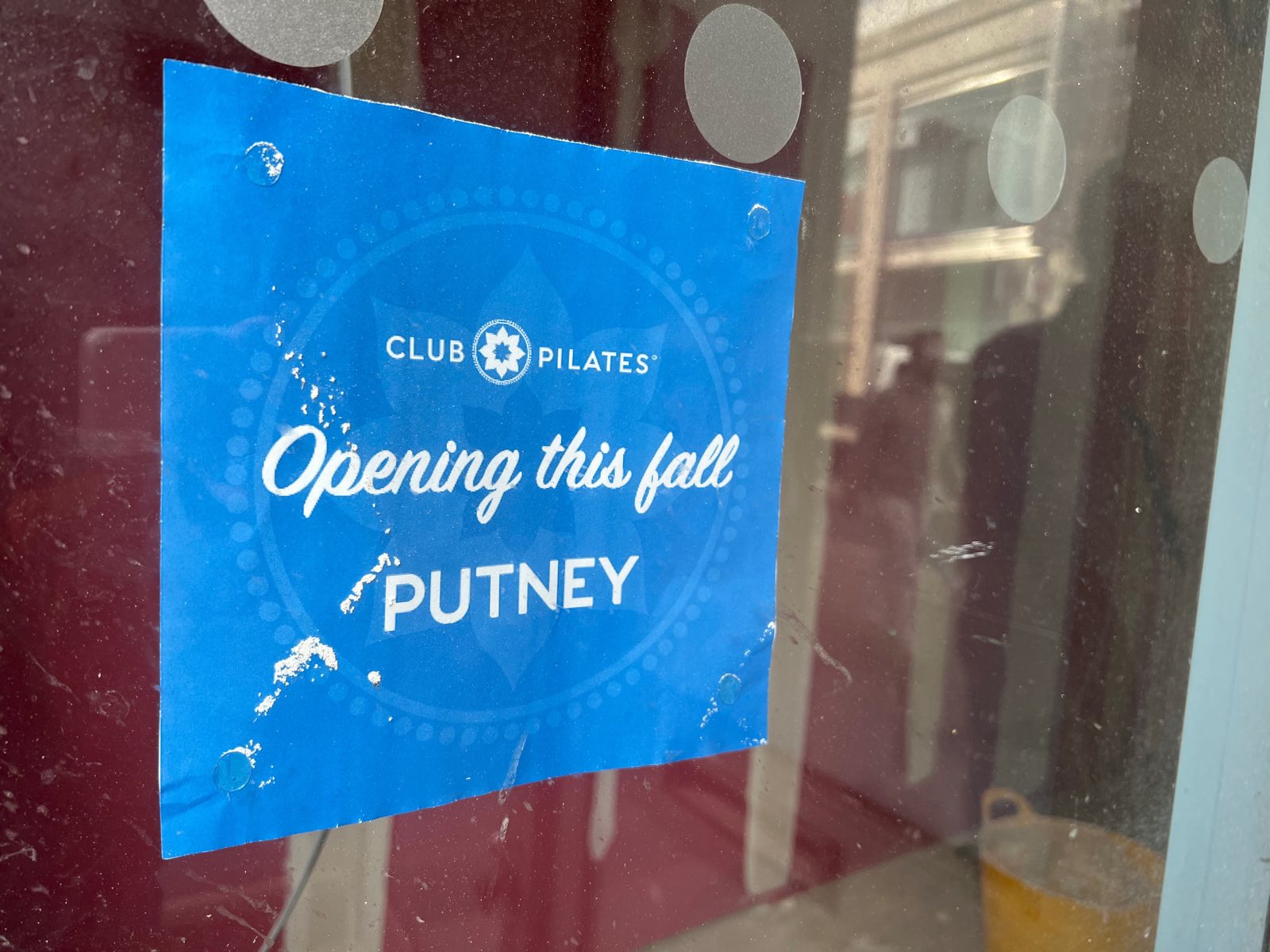Blue sticker in a shop window reading Club Pilates, Opening this fall, Putney