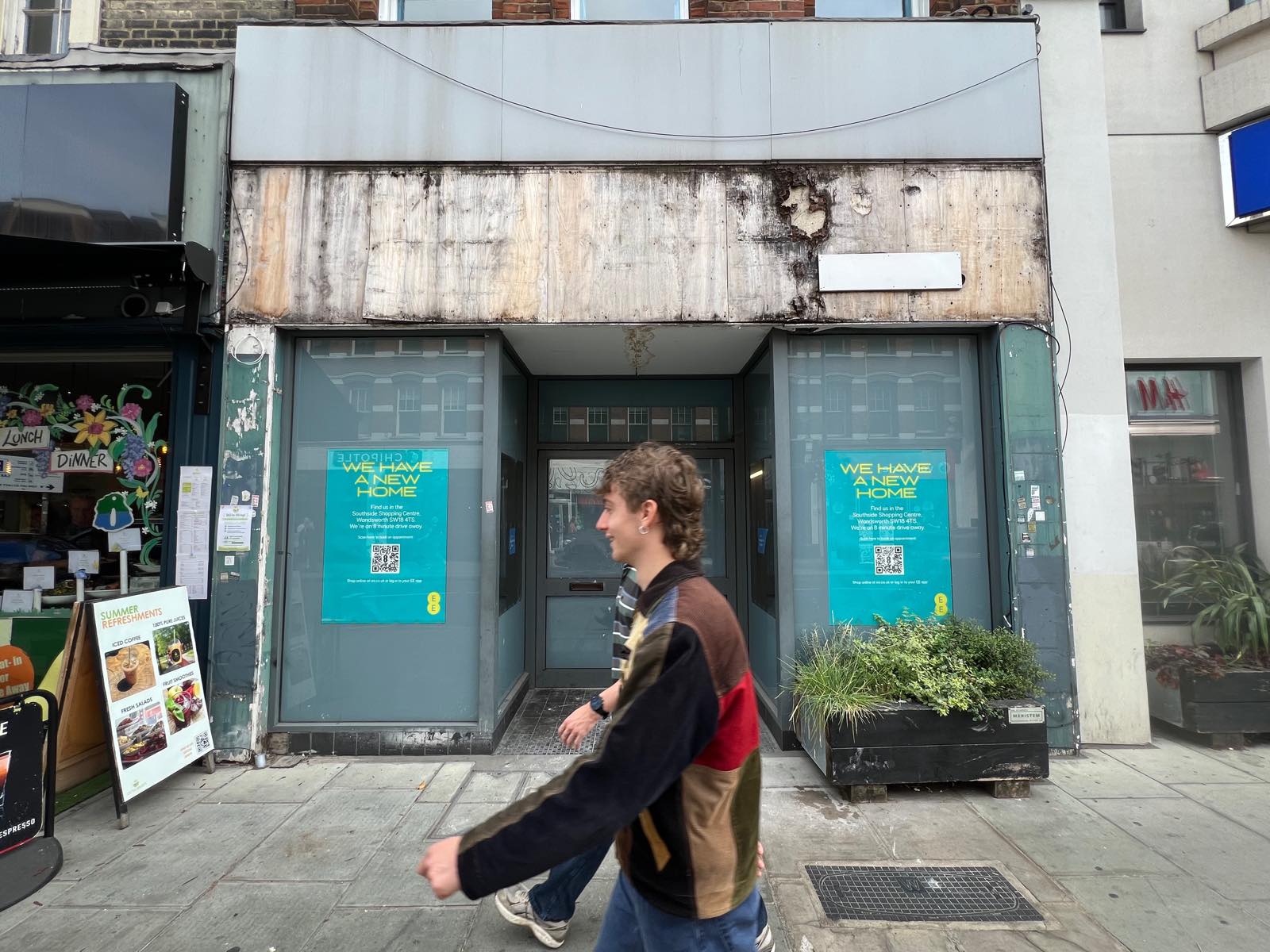 Boarded fascia of the former EE shop on Putney High Street with two teal EE posters in the windows reading We have a new home
