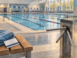 Empty public swimming pool with lane ropes, a membership card and folded towel on a poolside bench, and a closed turnstile in the foreground.