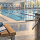 Empty public swimming pool with lane ropes, a membership card and folded towel on a poolside bench, and a closed turnstile in the foreground.