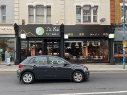 The Ta Ke Sushi frontage on Putney High Street in daylight, black fascia with Japanese lettering, KTV and izakaya signs over the pavement, the dining room visible through the glass and a car parked in front.