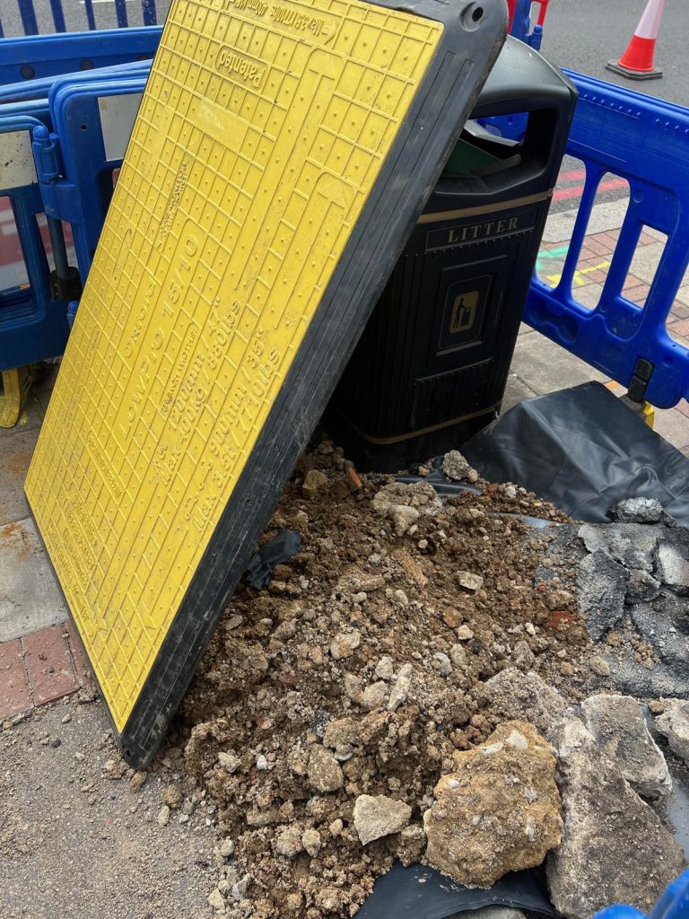 A yellow pedestrian trench cover propped against a litter bin beside a heap of spoil inside the fencing