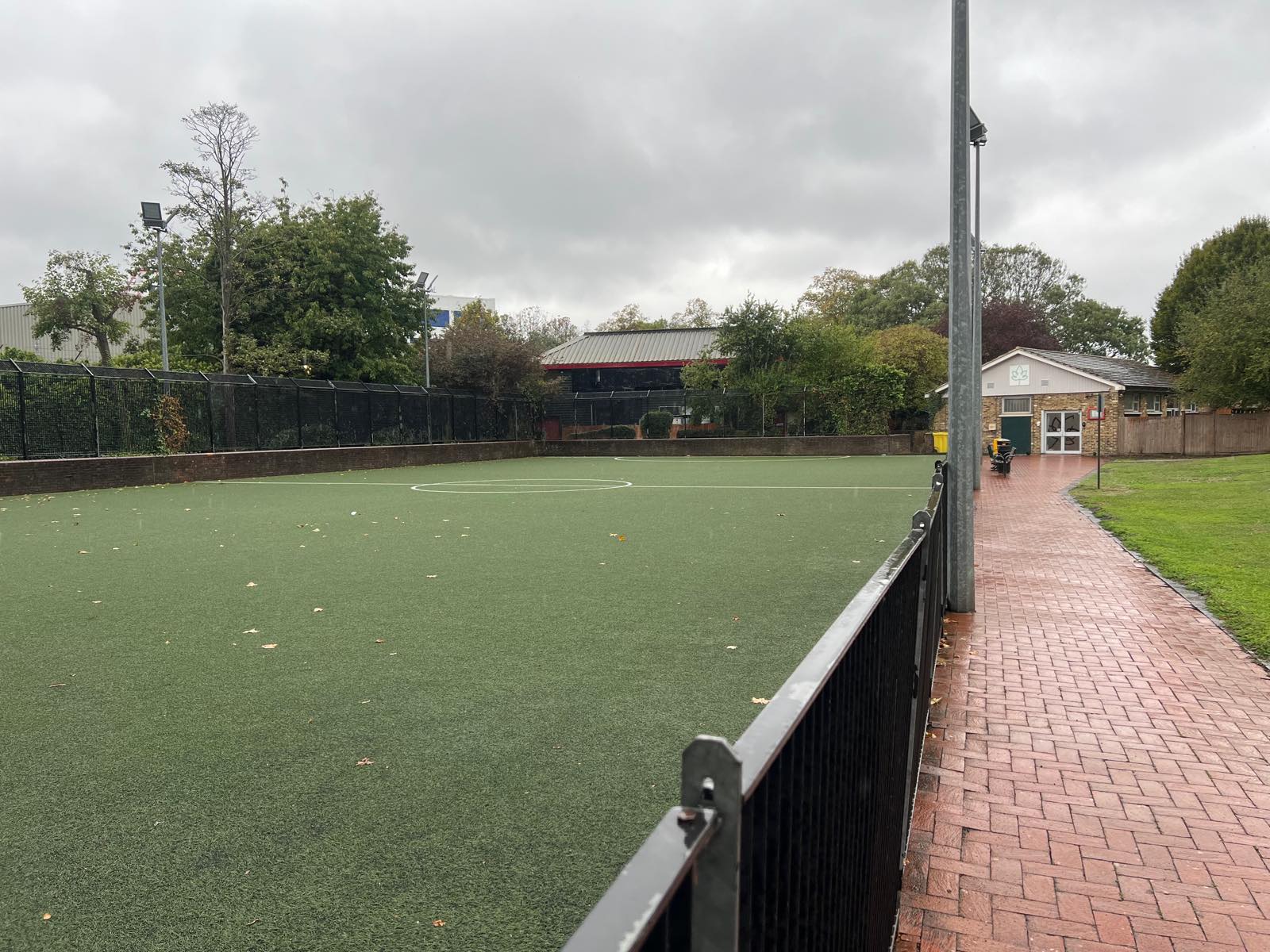 Pitch 5 at Wandle Recreation Centre seen from its far side, empty and wet, with the low brick wall and a floodlight column
